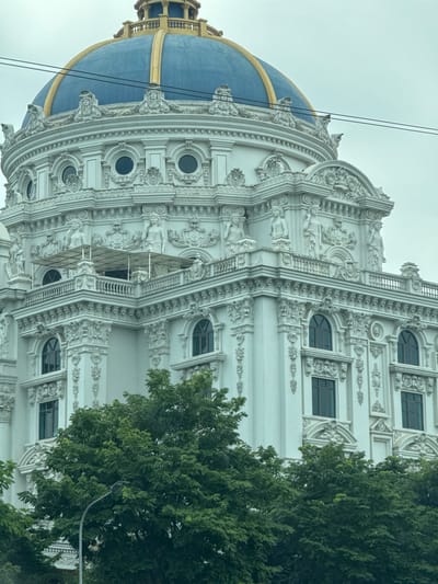 The image shows a portion of an ornate white building with a blue and gold dome, located in Hoa Lư Ward, Vietnam. The building's facade is decorated with intricate carvings, statues, and arched windows. There are trees in the foreground, and power lines are visible against the overcast sky. No people are visible in the image.