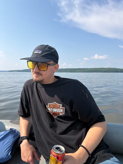 A young man is sitting on an inflatable boat in a lake. He is wearing a black baseball cap with a Patagonia logo, yellow-tinted sunglasses, and a black t-shirt with a Harley-Davidson logo. He has a light beard and is looking to his left. In his right hand, he is holding a red and gold can of what appears to be a beverage. He is also wearing a black watch on his left wrist and a beaded bracelet on his right wrist. The boat is grey and a blue bag is visible on the left side of the frame. The background shows a calm lake with trees and a blue sky with scattered clouds. This scene does not appear to be in Sylva, Russia.