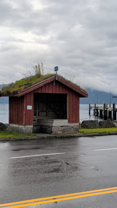 A rustic red wooden bus shelter with a grass-covered roof sits on a stone foundation next to a wet asphalt road. The shelter has an open front and a simple wooden bench inside. A blue bus stop sign is mounted on the roof's peak. In the background, a calm body of water is visible, leading to a wooden pier with several boats docked. Mountains partially shrouded in mist rise in the distance. The sky is overcast with heavy, grey clouds. The road in the foreground has white and yellow lane markings and is glistening from recent rain. This scene is located in the city of Halsa, Norway.