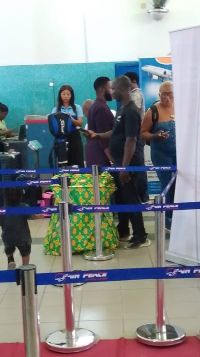 The image depicts an indoor scene, likely an airport check-in or boarding area, as suggested by the presence of queuing lines, a banner with an airplane logo and "AIR PEACE" branding, and passengers with luggage. In the foreground, a blue tape barrier with the "AIR PEACE" logo is visible, supported by metallic stanchions. Behind the barrier, a woman in a light blue top stands near a blue and black bag. To her right, a man in a dark purple shirt is speaking to another man in a black t-shirt. Further back, a woman in a denim jumpsuit with glasses is holding a phone. A child in a dark outfit is partially visible on the left, and a large, patterned green and yellow object, possibly luggage or a display, is situated in the middle ground. The environment appears to be a public space with tiled flooring and a light-colored wall. The scene is consistent with a location in Orerokpe, Nigeria, given the context of an airport.