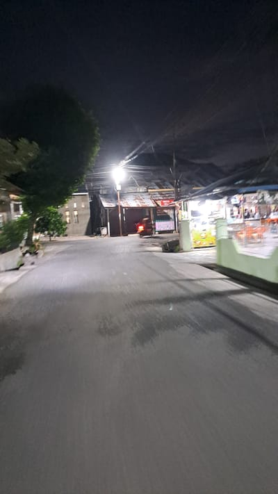 The image shows a street scene at night in Lhokawe Teungoh, Indonesia. A paved road runs through the center of the frame, appearing wet or reflective, possibly from recent rain. On the left side of the road, there is a large tree with dark green foliage and a building with a light-colored facade and dark windows. Further down the street on the left, another building with multiple windows is visible.
On the right side of the road, there are several structures with corrugated metal roofs. One structure appears to be a storefront or food stall with illuminated signs and displays, showcasing what might be food items or products. Orange chairs are visible within this illuminated area, suggesting an outdoor seating arrangement. A scooter with its taillight illuminated is parked in front of one of the buildings. Overhead, there are power lines and a bright street light illuminating the scene. The sky is dark, indicating it is nighttime. The overall impression is a dimly lit, possibly humid, urban or semi-urban environment at night.