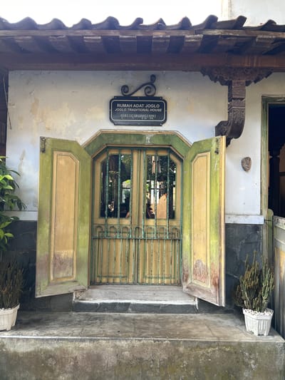 This media file shows an exterior view of a building, specifically a traditional Javanese house, known as a Joglo. The scene is oriented vertically, as if the photo was taken looking upwards or the camera was held sideways.

The main subject is a large, weathered wooden door painted in muted green and yellow. The door has decorative paneling and is fitted with a metal grate across its upper section, which reveals a glimpse of greenery and possibly other structures within. The door is partially open, with one of its panels swung outwards. Flanking the door are architectural elements consistent with Javanese traditional houses, including elaborately carved wooden brackets and a roof structure with tiled elements.

To the left of the door, a black plaque with white text is affixed to a textured, off-white wall. The visible text reads "RUMAH ADAT JOGLO TRADITIONAL HOUSE" followed by characters in what appears to be Javanese script. The wall itself shows signs of age with peeling paint.

On the right side of the door, a concrete structure forms part of the building's facade, and above it, a white planter holds dry, pine-like foliage. The overall setting appears to be a daytime scene, likely outdoors, given the lighting. The weathered appearance of the door and the old paint on the wall suggest a historic or traditional location. The location context is identified as Banguntapan, Indonesia, which aligns with the traditional Javanese architecture visible. No people are explicitly depicted.