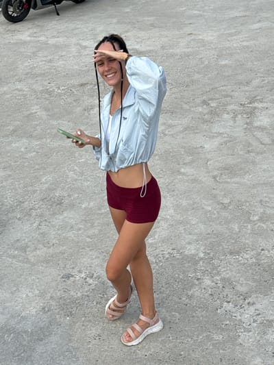 A young woman is standing on a concrete surface outdoors. She is wearing a light blue long-sleeved shirt, red shorts, and pink sandals. She is holding a green smartphone in her right hand and obscuring her face with her left hand. She is smiling. There is a black motorcycle partially visible on the left side of the image. The location is presumed to be somewhere within Lũng Phìn Commune, Vietnam, as specified in the request. The background consists of a textured concrete surface.