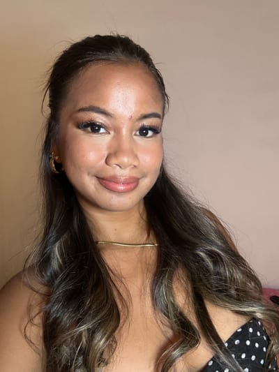 A woman with dark hair is facing the camera. She has light brown skin, dark eyes, and full lips. She is wearing a gold necklace and gold hoop earrings. She is wearing a black and white polka dot top. The background is a pale beige color. The image appears to be taken indoors. It is difficult to ascertain from the image whether or not this was taken in Celuk, Indonesia.