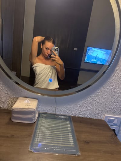 A woman is reflected in a circular mirror, taking a selfie with a smartphone. She is partially covered in a white towel. Behind her, the mirror reflects a wall-mounted television. To the right of the mirror, there is a tissue box and a menu card on a wooden surface. The location is a room in Sa Pa Ward, Vietnam.