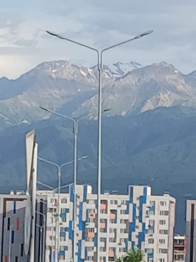 This is a vertically oriented, slightly blurry photograph of a modern, multi-story building with a blue and white facade, situated in a mountainous landscape. The building features numerous windows and balconies, with alternating sections of blue and white creating a geometric pattern across its exterior. In the foreground, a street lamp with multiple arms is visible, extending across the frame. Behind the building and street lamp, imposing mountains dominate the background, their rocky slopes partially covered in snow. The sky is a clear blue. The overall scene suggests an outdoor setting in a developed area bordering a natural, mountainous region. There is no clear indication of time of day or specific weather conditions beyond the clear sky. No people or discernible activities are captured in the image. No visible text is present. The location context provided is Іргелі ауылдық округі, Kazakhstan.