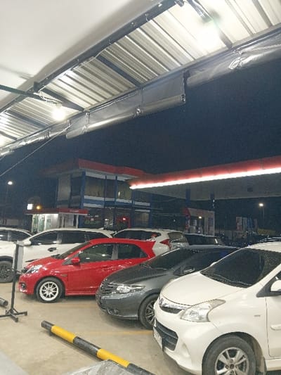 The image depicts a nighttime scene at an outdoor parking area in Seumadam, Indonesia, illuminated by artificial light. Overhead, a corrugated metal canopy structure with visible support beams and fluorescent light fixtures extends across the top of the frame. Below parts of the canopy, a dark, rolled fabric or material is attached. In the middle distance, a two-story building with blue-tinted windows, a flat roof, and red architectural accents is visible, likely a commercial or service station structure. A prominent horizontal red light strip runs along a section of the building or an adjacent structure.

In the foreground and middle ground, multiple cars are parked in rows on a concrete surface. From right to left, a white 5-door hatchback or MPV with a prominent front grille is partially visible. Adjacent to it is a dark grey 4-door sedan. To its left, a red 5-door hatchback with white multi-spoke wheels is parked. Further back, a white SUV and another white vehicle are partially visible. A black and yellow striped cylindrical parking barrier runs diagonally across the lower foreground of the image. No individuals are present, and no active events or interactions are captured within the static scene.