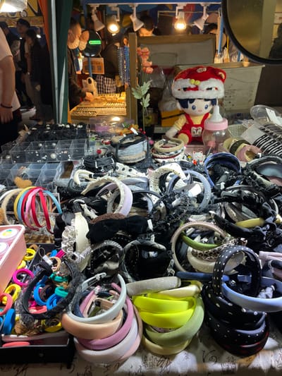 The image shows a street vendor stall in Hangzhou Shi, China, at coordinates 30.264 latitude and 120.156 longitude. The stall is selling a variety of headbands, hair ties, and other accessories. The stall is located in a busy area and the background shows people walking past. There is a plush doll wearing a red and white hat, a partially obscured white water bottle, and a number of black and white patterned headbands. The headbands are arranged on a table in front of the vendor, with the hair ties in a box to the left of the headbands.