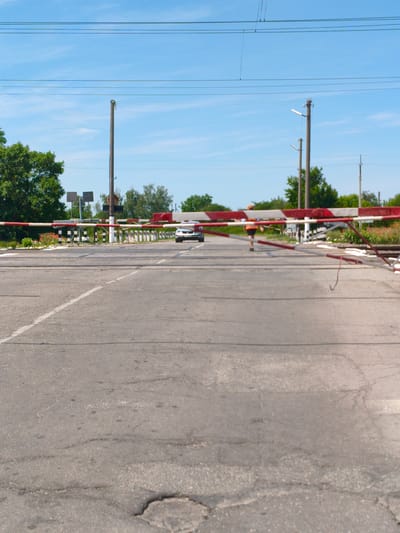 The image captures a railroad crossing in Hoholeve, Ukraine, on a sunny day with a clear blue sky. A road, showing signs of wear and tear with cracks and patches, crosses the railway tracks. A red and white striped barrier is lowered across the road, indicating an approaching or passing train. A silver car is visible on the other side of the barrier, stopped. A person wearing an orange vest and dark shorts is walking across the railway tracks, away from the car. Trees and greenery flank the sides of the road and tracks, and utility poles with overhead wires are present in the background. The scene depicts a typical moment at a guarded railroad crossing.