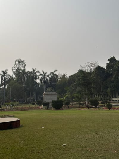 The image depicts an outdoor scene within Sayaji Baug (Kamati Baug) in Vadodara, India. An equestrian statue, identified as Maharaja Sayajirao Gaekwad III, stands prominently in the mid-ground. The statue, appearing to be crafted from a dark material consistent with bronze, features a male figure on horseback, positioned atop a multi-tiered, light-colored stone pedestal. The foreground is composed of a broad, green grass lawn, with a partial view of a low, circular, reddish-brown concrete border or planter in the lower-left. Surrounding the base of the statue are manicured garden beds, delineated by a low metal fence and incorporating trimmed green hedges and shrubs. The background consists of a dense array of mature trees, including numerous tall palm trees and other broadleaf species. The sky is a uniform, light gray, indicating an overcast or hazy condition, with a small, dark bird visible in flight in the upper-right quadrant. No human figures are present. Minor white debris is scattered on the grass in the foreground.