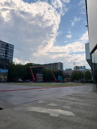 The image depicts a view of the Schouwburgplein in Rotterdam, Netherlands. In the foreground, a wet, paved area with puddles reflects the sky. Beyond this, a textured, metallic surface is visible, bordered by a red line. Further in the background, large, red, angular structures dominate the scene. These structures appear to be part of a modern art installation or a temporary stage setup. Behind these, a line of trees separates the foreground from a row of apartment buildings with numerous windows. In the distance, more buildings and a construction crane are visible under a cloudy sky. The overall impression is an urban outdoor public space.