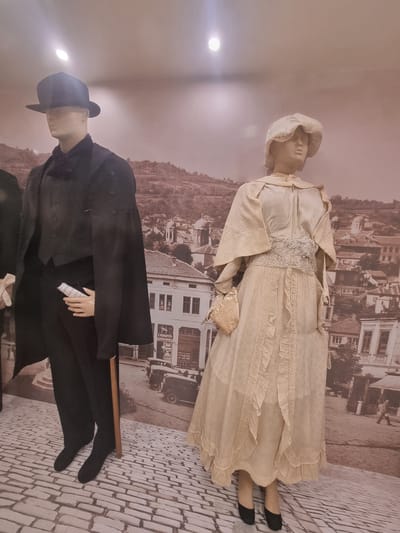The image shows two mannequins in historical attire, displayed against a backdrop of Gorna Oryahovitsa, Bulgaria. The female mannequin, on the right, wears a long, light-colored dress with ruffled details and a bonnet. She also carries a small purse. The male mannequin, on the left, is dressed in a dark suit with a hat and holds what appears to be a document. The backdrop shows a street scene, possibly a historical representation of the city. The setting appears to be an exhibit or museum. The floor has a cobblestone-patterned design that mimics the backdrop.