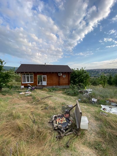A medium shot captures a rural scene on a cloudy day with a low angle. In the foreground, a stone fire pit with a grill is set up, containing meat and vegetables cooking. Scattered around the fire pit are pieces of wood. A wooden partition separates the fire pit from a white plastic container filled with water. To the left, a small wooden house with a dark roof and brown siding sits in the mid-ground. A white-framed window and an open door are visible on its front facade. Clothes are draped over a wooden structure near the house. To the right of the house, a white table with various items is set up amongst overgrown vegetation. In the background, a distant cityscape with buildings and hills is visible under a blue sky dotted with white clouds. The environment is characterized by dry grass and scattered trees.