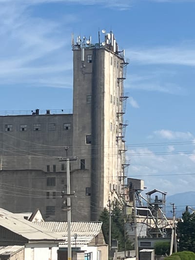 The image shows a tall, concrete industrial building, possibly a silo or a grain elevator, with a clear blue sky and some wispy clouds. The building is a dominant subject, featuring multiple levels and windows. Various antennas and communication equipment are mounted on top of the structure, suggesting it may also serve as a telecommunications tower. Power lines are visible crisscrossing the sky in front of the building.
To the right of the main building, there's a complex of metal structures, scaffolding, and smaller buildings, indicating ongoing construction, maintenance, or industrial operations. Trees and other vegetation are present, hinting at an outdoor setting. The lighting suggests it is daytime, likely in the morning or afternoon, with bright sunlight illuminating the scene.
The scene appears to be an industrial area, given the nature of the building and associated structures. There are no people visible in the image. The overall impression is one of functional, utilitarian architecture and industry. The location is identified as Balykchy, Kyrgyzstan.