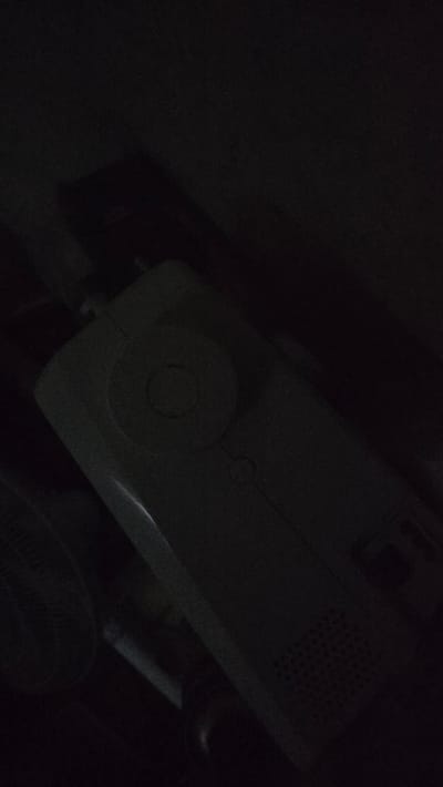 The image shows a dark, indoor scene with a white electronic device in the foreground. The device appears to be a portable power bank or similar electronic accessory, featuring a circular control dial on its top surface and a gridded ventilation area on its side. To the left of the device, a portion of a black fan with a circular grille is visible. The background is dark and indistinct, with hints of other objects, possibly furniture or boxes, also in shadow. The overall lighting is very low, suggesting the photo was taken in dim light conditions. There are no people or discernible actions depicted in the image. The scene is consistent with a personal space or storage area within a residence.