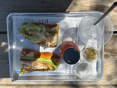 An overhead shot captures a casual outdoor meal served on a metal tray, resting on a rustic wooden table. The tray is lined with newspaper, upon which various food and drink items are arranged.
The main subjects are two halves of a sandwich, possibly made with a pretzel bun or baguette, filled with what appears to be pulled meat (like pork or chicken), fresh green lettuce, and topped with grated white cheese. The sandwiches show a golden-brown crust and a generous filling.
Accompanying the sandwiches are several side items in small clear plastic containers: one holds a serving of green olives, while another contains green herbs or spices. A red-orange bottle with a black cap, likely a beverage, sits between these containers and the sandwiches. In the bottom left, a clear plastic cup filled with ice and a metal straw indicates a cold drink.
The scene is brightly lit by natural sunlight, casting distinct shadows from the items onto the tray and the table, suggesting a sunny day. No people are visible, so no emotions are conveyed.
Visible text on the newspaper liner includes English headlines and articles, such as "Privacy Robbe:", "Just In", "Appetit", "liel emanuel or milo", and political news related to immigration and government funding. Notably, there is also Hebrew text, "יסמין והפצה בע"מ 08-6891887" (Yasmin and Distribution Ltd. 08-6891887), which aligns with the given location context of Eshkol Regional Council, Israel.