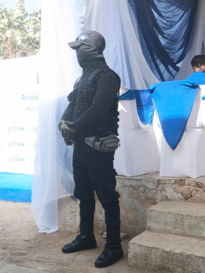 An individual dressed in black tactical gear stands guard at what appears to be an outdoor event in Tarauni, Nigeria. The person wears a black balaclava or face mask, a cap with reflective goggles pushed up, a tactical vest with numerous pockets, fingerless gloves, black trousers, and black boots. A grey utility pouch is fastened around their waist. They stand upright with hands clasped in front, facing left, indicating a watchful presence.

The event setup includes white sheer curtains on the left, partially revealing distant foliage and what seems to be electrical infrastructure. White chairs with blue fabric sashes are visible on the right, accompanied by a large blue fabric drape. A portion of another person wearing a blue t-shirt is seen behind the chairs. The ground consists of dirt and weathered concrete, with concrete steps leading up to the event space. The bright, natural light suggests a clear, sunny daytime. Banners in the background display the words "Retail," a partial "Activa," and "LACTOGROW" repeated several times, pointing to a product launch or promotional event for the Lactogrow brand. The scene depicts a secured outdoor gathering.