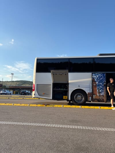 The image shows a large, white and black double-decker bus parked on an asphalt surface with yellow curbs. The bus has an image of London landmarks on its side. A man wearing a black shirt and shorts is visible inside the upper level of the bus. The sky is clear blue, and in the background, there are parked cars, a hill, and power lines. While the exact location within the Dimitrovgrad Municipality is not discernible from the image, it suggests a parking area near a roadside with views of surrounding landscape.