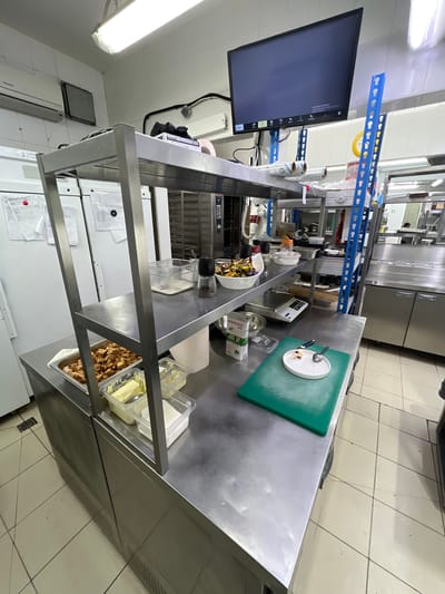 This is a wide-angle, eye-level photograph of a commercial kitchen environment. The scene is brightly lit, likely by fluorescent overhead lights, and captured in daylight. The camera is positioned at an angle that provides a clear view of a stainless steel preparation counter, shelves laden with various kitchen supplies, and a portion of industrial kitchen equipment.

Dominating the foreground is the stainless steel preparation counter. On it, a green cutting board is visible with a white plate containing a small amount of food residue and two spoons. Adjacent to this is a bowl filled with a mixture of food items, possibly a salad, and a pepper grinder. Further to the left on the counter are several clear plastic containers, one holding what appears to be butter, and another with white granulated sugar. A metal scale is also present on the counter.

Above the counter, a two-tiered stainless steel shelf unit holds a variety of items. The lower shelf contains a stainless steel tray filled with diced meat, likely chicken or pork, and more plastic containers. The upper shelf has additional containers, rolls of what appears to be plastic wrap or similar material, and a black object that could be a book or manual.

In the background, a large flat-screen monitor is mounted on a blue metal shelving unit. The monitor displays a dark screen with some text visible, possibly a control panel or menu. The blue shelving unit is tall and industrial, holding various items. Behind the shelves, industrial kitchen appliances are visible, including what look like ovens or proofers. To the left of the preparation counter, large white appliances, possibly refrigerators or freezers, are partially visible. The walls of the kitchen are tiled in white, and the floor is tiled in a light beige color with dark grout lines.

There are no people clearly visible in the frame. The overall impression is one of an organized, active food preparation space.

The location is identified as Plast univska, Russia.