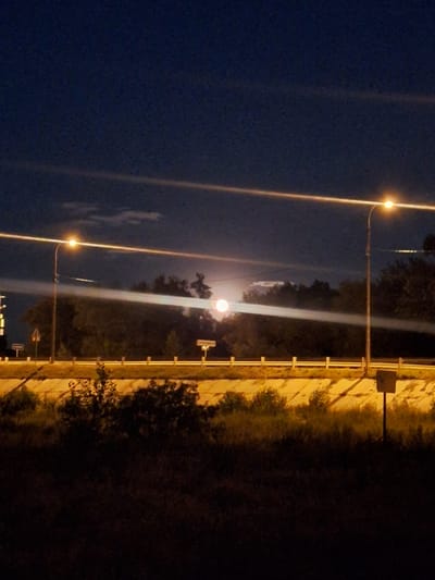 The image captures a nighttime scene on the outskirts of Стеженский, Russia, likely near a road. A full moon, appearing as a bright orb, is partially obscured by scattered clouds in the dark blue sky. Two streetlights are visible, casting distinct beams of light across the frame. The one on the left illuminates a section of the sky and a tree line. The streetlight on the right emits a strong beam that extends horizontally, illuminating clouds and a portion of the landscape. In the foreground, a dark, grassy area suggests undeveloped land. Beyond this, a raised concrete barrier runs horizontally, separating the road from the foreground. Behind the barrier, a dense line of trees forms a dark backdrop. The overall atmosphere is one of quiet twilight, illuminated by artificial light and the moon. There are no people or discernible actions taking place.