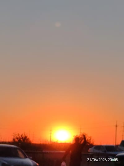 A low-angle, slightly blurry photograph captures a sunset over a suburban or rural landscape, possibly in or near Козет, Russia. The sky transitions from a light gray-blue at the top to a vibrant orange-yellow near the horizon. The sun, a bright white orb, is low in the sky, casting a warm glow. A faint, pale circle is visible in the upper left portion of the sky, possibly the moon or a lens flare.
In the foreground, silhouetted figures and vehicles are visible. On the left, the front portion of a dark-colored car is present. In the center, a person is standing with their arms raised, appearing to be celebrating or interacting with someone. To the right of this person, parts of other vehicles are visible. The environment appears to be an open area with some sparse vegetation and possibly utility poles in the background. A timestamp "21/06/2026 20:05" is overlaid on the bottom right of the image, indicating the date and time the photograph was taken.
