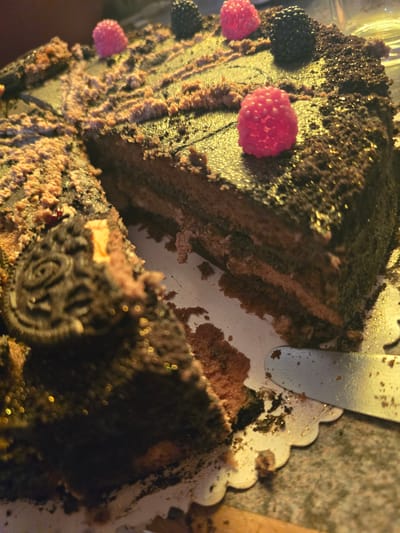 The image shows a partially cut chocolate cake. It is decorated with raspberries and what appear to be blackberries or chocolate decorations resembling blackberries. A piece of the cake has been removed, revealing the layers. A silver serving utensil is visible near the cut edge of the cake. The cake sits on a white, scalloped-edged cake board. There is no indication of the location being Zirndorf, Germany. The image focuses on the cake; there are no people or specific environmental clues.