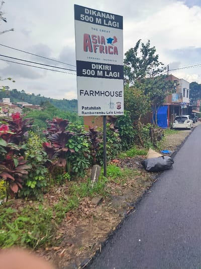 A sign post stands on the side of a paved road in Lembang, Indonesia (latitude: -6.826, longitude: 107.608). The sign reads "Dikanan 500 M Lagi," "The Great Asia Africa, Lembang - Indonesia," "Dikiri 500 M Lagi," and "Farmhouse. Patuhilah Rambu-rambu Lalu Lintas," which translates to "Right 500 M Ahead," "The Great Asia Africa, Lembang - Indonesia," "Left 500 M Ahead," and "Farmhouse. Obey Traffic Signs." There are lush green bushes with red foliage to the left of the sign, and the road is relatively empty. A black garbage bag is on the shoulder of the road near the sign post. A small blue tarp hangs on a structure to the right of the road. In the distance, a small gray car is driving down the road. The sky is overcast with light gray clouds.