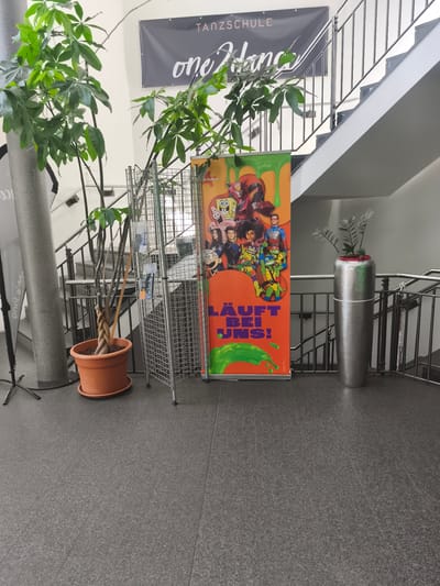The image shows an indoor scene, likely a school or community center hallway, with a staircase in the background. To the left, a tall potted plant with braided stems stands in an orange pot. Beside it, a metal display rack holds various items. In the center of the image, an orange banner featuring characters from Nickelodeon shows like SpongeBob SquarePants and the Teenage Mutant Ninja Turtles, with the text "LÄUFT BEI UNS!" (It's on for us!) in large, bold letters. To the right, a tall silver cylindrical planter holds a small green plant. A dark gray floor covers the ground. A banner reading "TANKSCHULE one dance" hangs above the staircase. This scene is consistent with the entrance area of the Tanzschule ONE DANCE in Niederkassel, Germany, located at Mondorfer Straße 12, 53859 Niederkassel.