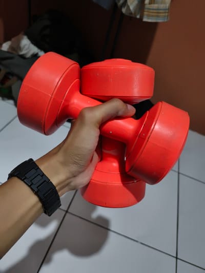 A person holds two red plastic dumbbells stacked atop one another. The person wears a black watch. The image is taken indoors on a tile floor, potentially in a home gym setting. Part of the wall is peach colored. Some items are placed in the background. It is impossible to determine if this image was taken in Pacet, Indonesia.