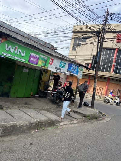 The scene is a street corner in Kecamatan Margahayu, Indonesia (latitude: -6.971, longitude: 107.574). A woman wearing a hijab and light blue jeans walks on a sidewalk towards the camera. She is wearing a black sweater with white stripes, black shoes, and a white hijab. She is walking past a green building with a sign for "WIN SWEET AROMATIC". To her right, another person is walking towards the camera. He is wearing a black jacket and dark pants, carrying a black bag over his shoulder. He is passing a black motorcycle parked on the side of the road. In the background, a second motorcycle is parked on the other side of the road. The road is empty. The building behind the sidewalk has a large window facing the street. There are several power lines running across the image. The image is taken from the street level. The weather appears to be cloudy. The scene is located in a city area, and the building is likely a commercial property.