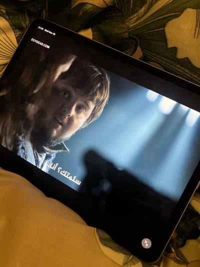 An iPad displays a scene from a television show or movie. The scene features a male actor with light brown hair and stubble. He appears to be lying down, looking slightly upward with an expression of concern. Arabic subtitles are visible on the lower left side of the screen. In the upper right corner of the screen, there is an icon resembling two opposing arrows within a circle. The iPad rests on a surface with a floral patterned fabric, which may be a blanket or pillow.