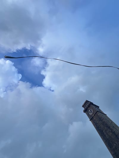The image is an upward-facing shot of a cloudy sky with a stone clock tower visible in the upper right corner and a dark, thin object hanging from the top left of the frame. The clock tower appears to be made of weathered stone blocks and has at least two clock faces visible, each with Roman numerals. The sky is a mix of pale blue and grey clouds, suggesting overcast or partly cloudy weather. The dark object in the foreground is out of focus and appears to be a cable or rope, possibly for a flag or another suspended item. The perspective suggests the photograph was taken from below, looking up at the tower and the sky. The overall impression is one of a potentially dramatic or imposing structure against a dynamic sky. There is no visible text in the image. The time of day is not definitively discernible, but the light quality could suggest late afternoon or an overcast day.