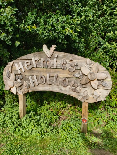 The image shows a wooden sign in the city of Rugby, United Kingdom, with the words "hermits hollow" carved into it. The sign is decorated with wooden cutouts of leaves, flowers, and butterflies. It is set against a backdrop of green trees and foliage. The sign is supported by two wooden posts, and a small circular sign with a red paw print and the words "No dogs" is attached to the right post. The ground around the sign is covered in grass and green plants.