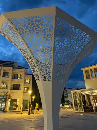 The image depicts an outdoor scene at dusk or twilight in Kumbor, Montenegro. A large, white, geometric structure with a triangular cut-out pattern dominates the foreground, curving upwards. Behind the structure, modern buildings with multiple floors and illuminated windows are visible. A restaurant with a glass facade and outdoor seating is also present.
Several people are scattered on the paved walkway in the middle ground. Some are standing, while others appear to be with a stroller, suggesting a casual evening stroll or gathering. The lighting suggests it is evening, with artificial lights from lamps and buildings illuminating the area. The sky is a deep blue with some clouds, indicating the transition from day to night.
There are no overtly strong emotions conveyed by the people, appearing relaxed in the environment. The scene suggests a public space, likely a promenade or a town square, designed for leisure and social interaction.
Visible text includes "PORTOM" on a white banner or sign on the right side of the image. The overall impression is one of a modern, well-lit urban or resort area at the end of the day.