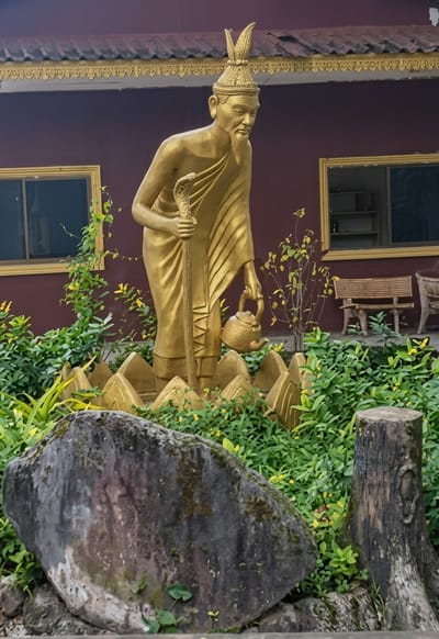 A golden statue of a robed figure stands in front of a maroon building with gold trim. The figure wears a tall headdress and holds a staff topped with a snake, and a small teapot in its other hand. The statue is surrounded by lush green foliage and yellow flowers. In the background, there are windows, benches, and a wooden stump. This scene is located in Sihanoukville, Cambodia, outside the Wat Leu temple.