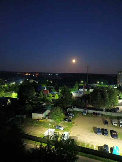 This is a nighttime aerial photograph taken in Alexandrov, Russia. The scene depicts a semi-urban or suburban landscape under a dark blue, clear night sky with a bright full moon visible. The moon is the most prominent celestial object, casting a soft light over the area.
The landscape is characterized by a mix of residential buildings, industrial structures, and dense vegetation. In the foreground, there is a dark, unpaved area with several parked cars, illuminated by artificial lights. These lights also highlight a fence and some grassy patches. Further back, there are various buildings, some appearing to be houses with lit windows, and others that look like small industrial or utility buildings. A tall tower, possibly a communication tower or crane, stands prominently in the middle ground.
The overall atmosphere is serene and quiet, typical of a rural or suburban area at night. The artificial lighting creates pools of brightness amidst the darkness, defining the spaces and structures. The presence of cars suggests a populated area, likely a residential community or a place of work. The time of day is clearly night, as indicated by the dark sky and artificial illumination. No people are directly visible in the image, but their presence is implied by the parked vehicles and lit homes. The weather appears to be clear, as the moon is vividly visible and there are no signs of rain or fog.