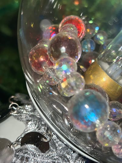 The image provides a close-up, high-angle perspective of a decorative arrangement featuring numerous translucent and iridescent spheres within a clear glass vessel. The main subjects are sparkling, spherical beads or ornaments.

The beads display a range of colors and light effects. Many are clear with shimmering, rainbow-like iridescence, scattering light into various hues. Some exhibit more pronounced colors, including vibrant reds and oranges in the lower left, and a dominant, larger sphere in the upper right that glows with distinct blue and green iridescent tones. These beads are reflective, showing bright specular highlights.

Within the cluster of beads, a cylindrical object with a matte gold metallic finish is partially visible. Close examination of a cropped section reveals faint, embossed details on its surface, possibly stylized text like "EA" or a decorative pattern, though it is largely indistinct.

The container holding these items is a clear glass vessel with a curved rim, visible along the right edge of the image. To the right and slightly below the glass rim, outside the vessel, there is a tangled mass of shiny, reflective silver material, resembling fine metallic tinsel or an intricate chain. Attached to this silver material are several small, flat, irregularly shaped silver metallic charms or sequins, connected by small jump rings.

The background is softly blurred into dark green tones, suggesting foliage, possibly from a Christmas tree or other decorative greenery, creating a shallow depth of field that highlights the intricate details of the foreground. No specific activity or event is depicted; the image captures a decorative still life.