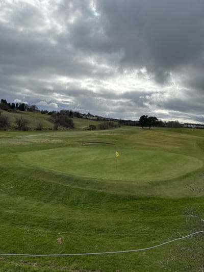 The image shows a golf course in Dunfermline, United Kingdom. The foreground consists of a large, green grassy area with a yellow flag marking a hole. The background shows trees, a grassy hill, and what appears to be buildings in the distance. The sky is overcast with grey clouds. A grey electrical cable runs from the upper right corner into the image. There is no evidence of people.