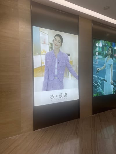 Two large digital billboards are displayed in a modern, indoor commercial space with marble-tiled walls and flooring. The billboard on the left features a woman wearing a lavender tweed suit and a white top, posing with a soft smile. The billboard on the right shows a woman in a white dress, holding onto a bicycle with a basket, and appearing to smoke a cigarette, with Chinese text that translates to "Design, Style, Purity" above her. The context suggests these billboards are likely part of a retail establishment or shopping mall in Liandu District, China, promoting fashion or lifestyle brands.