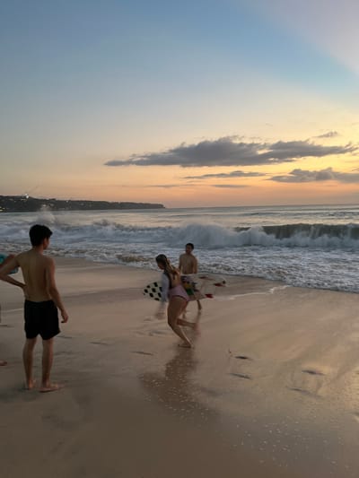 This media file depicts a beach scene with several individuals. The primary subjects are three people on the sand near the ocean. One person, a male, is standing and facing away from the camera, wearing dark swim trunks. Another person, a female, is lying on the wet sand, wearing a purple swimsuit, and a checkered towel is beside her. A third person, a male, is partially visible behind her, also on the sand. Further out, a large wave is breaking, with foamy water reaching the shore. The sky above displays a gradient of colors, transitioning from a pale blue at the top to warm orange and yellow hues near the horizon, indicating it is either sunrise or sunset. Several clouds are present in the sky, some appearing dark and dramatic. The sand is wet and reflective in places, with small puddles and ripples from the receding waves. The overall setting is a beach with the ocean and sky as prominent features. The activity appears to be people relaxing and enjoying the beach environment. The time of day is likely dawn or dusk due to the lighting and sky colors. The location context provided is Kuta Selatan, Indonesia, which is known for its beaches. No visible text is present in the media file. The mood appears to be tranquil and relaxed.
