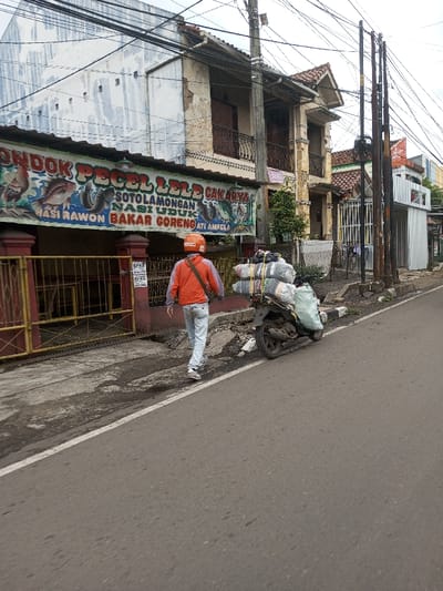 The scene is depicted on a street in what the prompt identifies as Kodarapalli, India, though visual cues such as signage for "Pecel Lele Cak Arya" and "Sotolamongan Nasi Uduk" suggest an Indonesian context. One individual, seen from behind, walks from right to left on a concrete sidewalk, wearing an orange long-sleeved jacket, light blue jeans, white athletic shoes, and an orange helmet with a white logo. A dark messenger bag is worn across their body.
To the individual's right, a dark-colored scooter-style motorcycle is parked on the curb, heavily loaded with multiple light-colored, bundled packages secured by various straps, and an additional large, light green bag attached to the rear.
The left side of the street features a single-story establishment with a prominent, colorful banner sign advertising "PONDOK PECEL LELE CAK ARYA," "SOTOLAMONGAN NASI UDUK," and "BAKAR GORENG ATI AMPELA," adorned with illustrations of fish and chicken. Behind this, a two-story building with a light blue/grey corrugated metal upper facade is visible. Adjacent to it, another multi-story building with a beige plaster facade, featuring balconies with dark railings and decorative columns, exhibits signs of wear. Further right, additional structures, including a narrow white building with vertical corrugated metal siding, line the street. Numerous dark utility poles are erected along the right side of the road, supporting a dense network of overhead wires. The foreground consists of an asphalt road with a partial white lane marking. The sky is overcast.