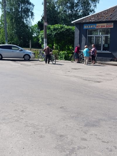 A light-colored sedan is parked to the left of a dark-colored building with a sloped roof. The building has a sign with blue and yellow lettering that reads "МАГАЗИН ВІЗИТ ВСЕ ДЛЯ ДОМУ" in Ukrainian. Three people are standing in front of the building, near two bicycles. One person is wearing a brown shirt and black pants, holding onto a bicycle. Another person is wearing a red long-sleeved shirt and dark shorts, and a third person is wearing a blue t-shirt and light-colored pants. A fourth person, wearing a patterned shirt, is standing further to the right. The ground is paved with asphalt, and trees are visible in the background. This scene is located in Luka, Ukraine.