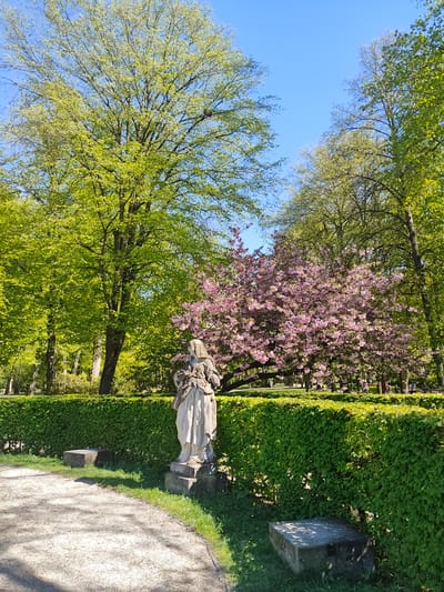 The image shows a statue in the Hofgarten, a park in Bayreuth, Germany. The statue is positioned along a gravel pathway bordered by a manicured green hedge. In the background, trees with green and pink blossoms are visible against a clear blue sky.