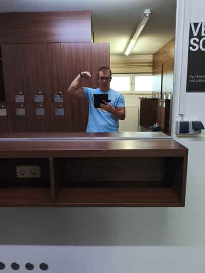 The image shows a man taking a mirror selfie in a locker room. He is wearing a blue t-shirt and flexing his bicep. The locker room appears to be in a gym or spa, with rows of wooden lockers visible in the background. There is a shelf in the foreground with two electrical outlets. The lighting in the room is fluorescent. A sign with German text is visible on the right wall, with the words "VERFÜHRERISCH UND SCHÖN" visible. The city of Pegnitz, Germany is not depicted in this image.