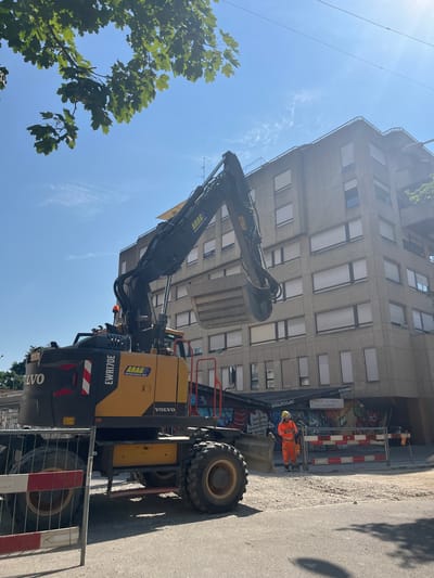 The image shows a construction scene on a sunny day in Geneva, Switzerland. The main subjects are an excavator and a construction worker. The excavator is yellow and black, with "VOLVO" and "EWR170E" clearly visible on its side. The excavator's arm is raised, and its bucket is in focus. A construction worker, dressed in an orange high-visibility suit and helmet, stands near the excavator, holding a shovel.

The setting is an urban environment, with a multi-story building in the background. The building has a concrete facade and numerous windows, some with blinds. Graffiti art is visible on the wall of the building behind a construction barrier. The ground is paved with asphalt, and there are loose stones and debris present, indicating ongoing construction work. A metal safety barrier with red and white markings is also in the foreground.

The time of day appears to be midday, given the bright sunlight and the shadows cast. The weather is clear and sunny, with a blue sky and a few wispy clouds. The overall impression is one of an active construction site. The image is oriented vertically.