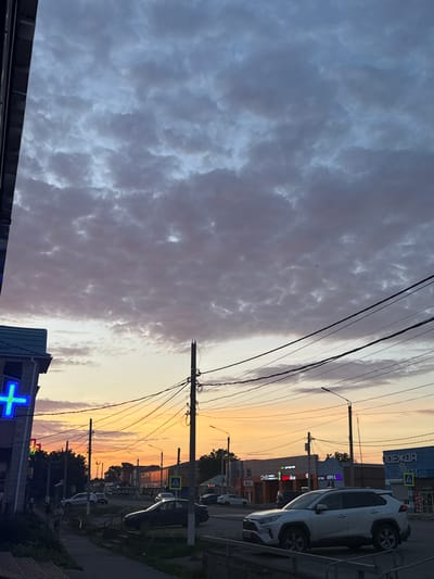 The image depicts a street scene in Yegorlykskaya, Russia, during a sunset. The sky is filled with a mix of dark and light clouds, suggesting a transition in weather or time of day. Utility poles with numerous wires dominate the foreground and mid-ground, stretching across the frame and obscuring parts of the buildings and sky.

Several buildings line the street, some featuring illuminated signs. Visible text includes "ОДЕЖДА" (Clothing) on a blue sign, and other signs with Cyrillic script that are partially obscured or too blurry to read clearly. One sign appears to be for a pharmacy, indicated by a glowing blue cross symbol. A white SUV is parked prominently on the right side of the street, facing away from the viewer, and another smaller, dark vehicle is visible further down the road.

The setting is a typical urban or suburban street with a paved road and sidewalks. The warm hues of the sunset cast a soft glow on the scene, contrasting with the darker clouds. The overall mood is somewhat muted due to the time of day and overcast sky, but the illuminated signs and the sunset provide a sense of activity and visual interest. There are no discernible people in the image. The time of day is clearly sunset, and the weather appears to be partly cloudy.