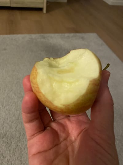 A hand holds a partially eaten apple. The apple is red and yellow, with bite marks revealing its white flesh. The hand is lightly colored and appears to be an adult's. The background is blurred, showing a wooden floor and a grey carpet. The image is taken indoors.