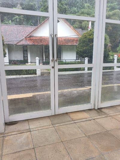 The image depicts an exterior view from inside a building in Munyati, Zimbabwe, looking through a set of white-framed glass doors or windows. The foreground consists of light brown tiled flooring. The glass panels are smudged and show visible streaks and droplets of falling rain. Beyond the glass, a wet, dark asphalt road with a yellow painted line is visible. Across the road, a black metal fence with white pillars separates the road from a property. Behind the fence, a white building with a distinctive reddish-brown tiled roof and multiple rectangular windows is situated. Green shrubs and trees are present in front of and surrounding the building. Dense green foliage and a white flag with a red emblem are partially visible further in the background, suggesting a heavily vegetated environment. No individuals are present in the scene. The weather conditions indicate active rainfall.