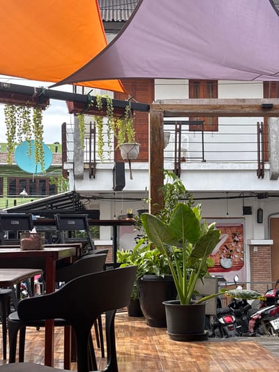 An outdoor dining area in Tambon Ko Tao, Thailand, is depicted under an overcast sky. The foreground features an arrangement of dark-colored tables and chairs on a reddish-brown patterned tiled floor. Overhead, orange and purple triangular shade sails are stretched, supported by dark metal beams. These beams also support wooden planters with trailing green foliage and white macrame hanging baskets containing potted plants. A square tissue box is present on one table.

In the midground, several large green potted plants, including an Alocasia species, are arranged on the tiled floor. Beyond the seating area, a multi-story building is visible. Its lower section features a white facade, while the upper story has horizontal wooden paneling and brown-framed windows. A balcony on the upper level is equipped with black horizontal railings, a white plastic chair, and a concrete bust. To the left, a building with green siding and dark windows is partially visible, with a light blue satellite dish mounted on its exterior. Below the main building's balcony, a banner with a graphic of hands and flowers is displayed on a wall. Several motorcycles and scooters are parked to the right on the tiled surface, partially obscuring a second graphic. No individuals are present in the scene, and no actions or interactions are occurring.