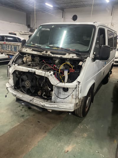 In Panyu District, China, a white van is parked in an industrial garage. The van's front end is dismantled, revealing the engine and its components. Various wires and parts are exposed, suggesting ongoing repair or maintenance work. The van is positioned on a green concrete floor, with some dirt and debris scattered around. Other vehicles and parts of the garage structure, including fluorescent lighting, are visible in the background.